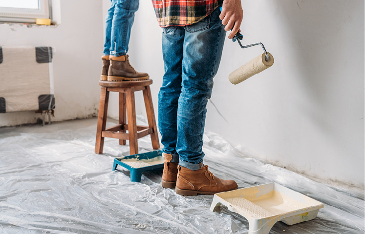 two people painting a wall, one standing with a paint roller and one on a stool