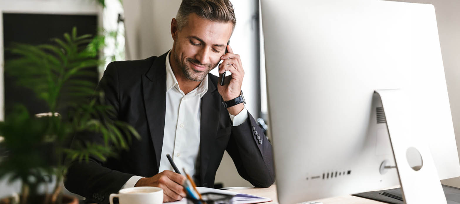 Man on phone writes notes in a notebook at his desk