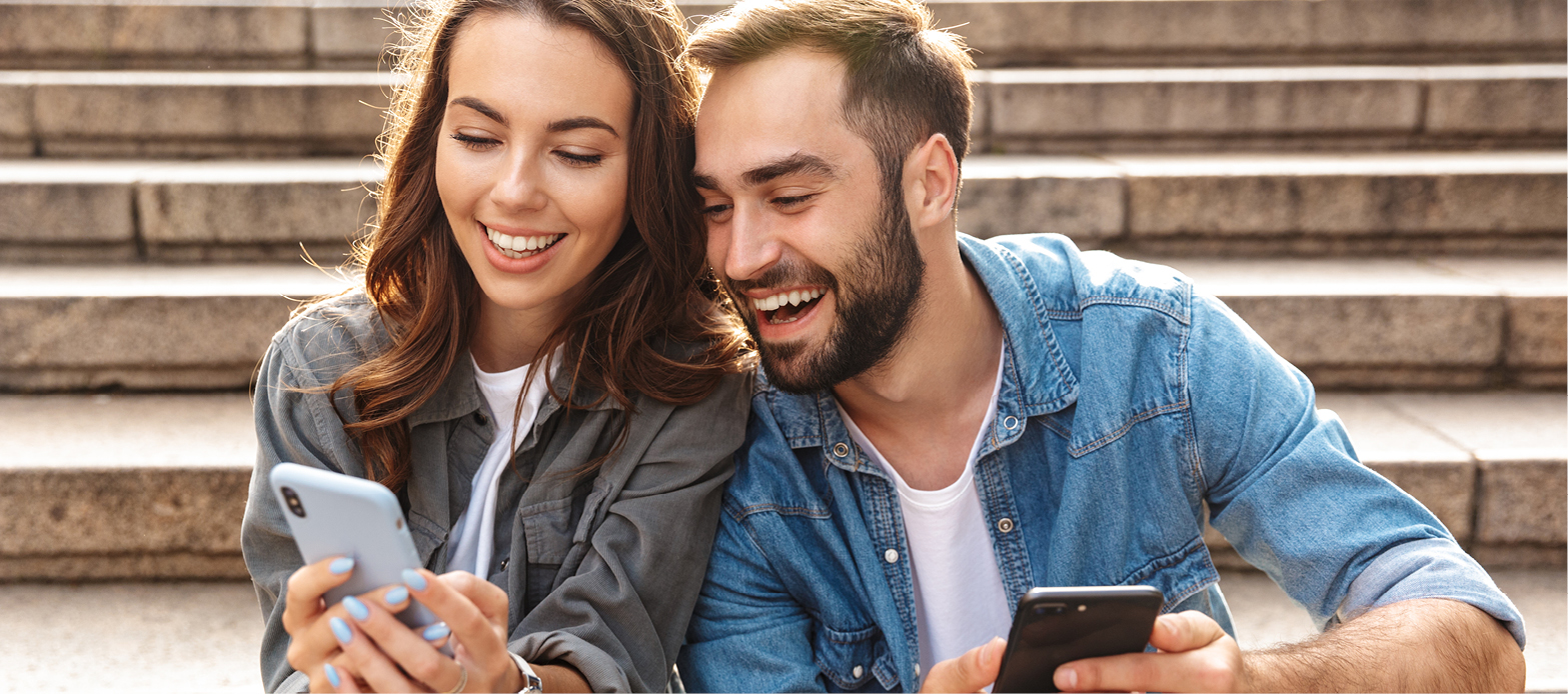 Male and female sitting on steps smiling and laughing at cell phones.
